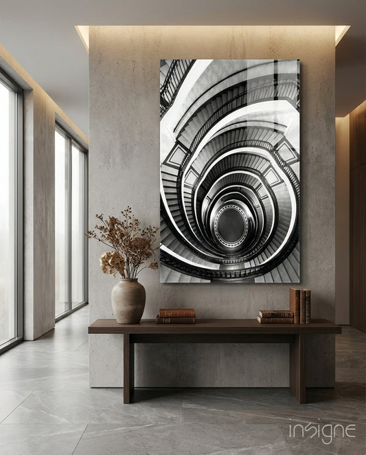 Monochrome spiral staircase glass wall art displayed above wooden console table in minimalist interior with concrete walls and dried flowers.