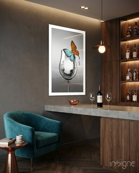 Framed white-bordered wall art depicting a wine glass with blue and orange butterflies, displayed above a bar counter with bottles.