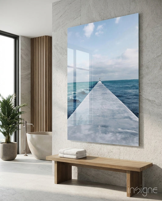 Large glass wall art depicting a white pier extending into turquoise ocean under cloudy sky, installed in modern bathroom above wooden bench.