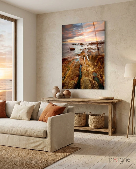 Golden Rocky Coast At Sunset glass wall art displayed above wooden console table in coastal-themed living room with cream sofa and ocean view window.