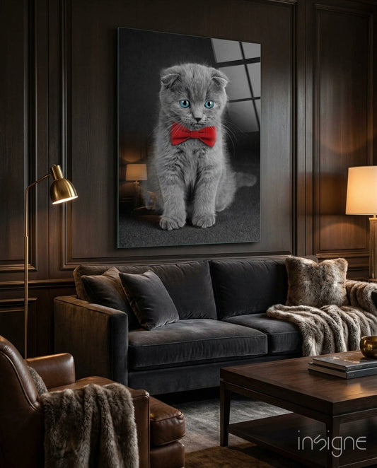 Gray Scottish Fold kitten wearing red bow tie displayed on glass wall art above black sofa in luxury living room.