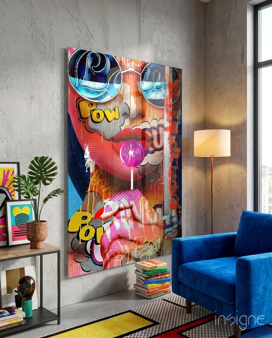 Colorful pop art glass wall panel featuring neon pink lips, blue swirls, yellow "pow" text, and comic-style elements displayed in modern living room with blue velvet chair.