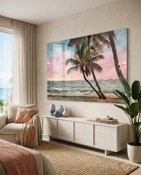 Beach scene glass wall art depicting leaning palm trees at pink sunset, displayed above white credenza in coastal bedroom.