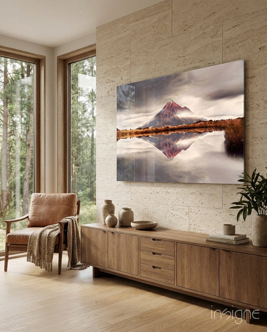 Mounted on a travertine wall above a wooden credenza, glass wall art depicting a red-peaked mountain reflected in still water below a dramatic sky.