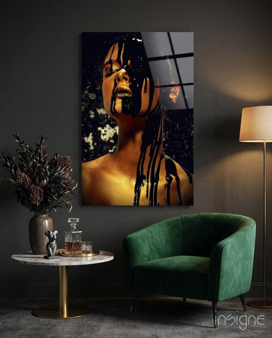 Stain glass wall art depicting a woman's face with golden skin and black dripping paint effect, displayed above a green velvet chair in a styled interior setting.