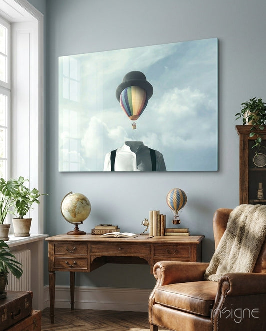 Glass wall art depicting a figure with a hot air balloon replacing their head, displayed above a wooden desk in a styled interior room.