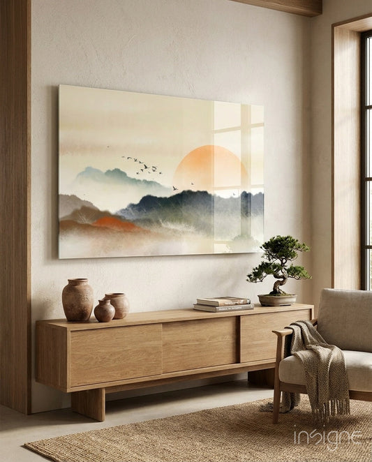 Abstract landscape glass wall art featuring misty mountains, orange sun, and flying birds displayed above wooden console in minimalist interior.
