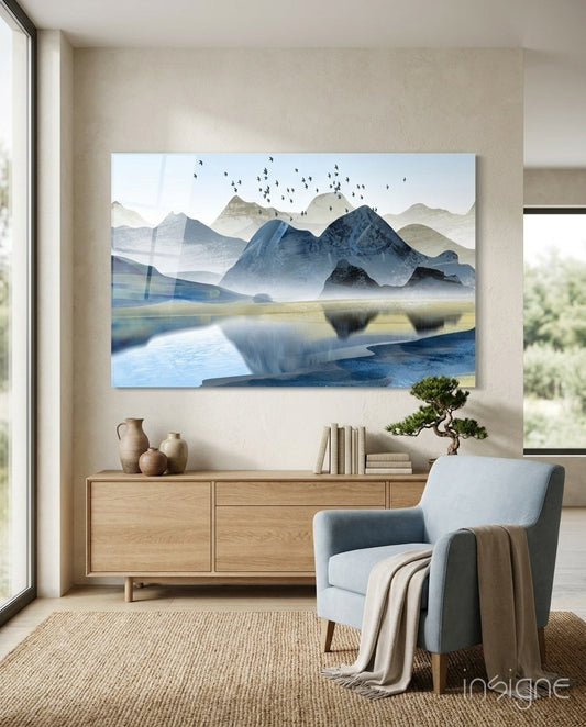 Glass wall art depicting misty blue mountains, a reflective lake, and flying birds, displayed above a wooden dresser in a modern living room.