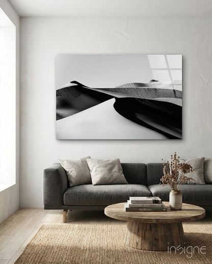 Black and white glass wall art depicting abstract curved sand dunes displayed above a gray sofa in a minimalist living room.