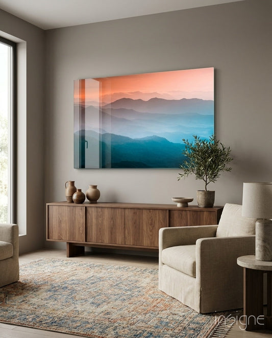 Orange and blue gradient mountain landscape glass art hanging above wooden credenza in modern living room with beige chairs and potted plant.