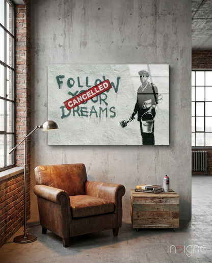 Banksy-style glass art depicting stenciled figure with "Follow Your Dreams" text crossed by red "Cancelled" banner, displayed on concrete wall above leather chair.