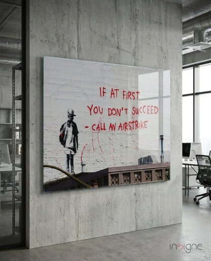 Glass art panel mounted on concrete wall in modern office, featuring stenciled child figure and red text reading "If at first you don't succeed - call an airstrike.