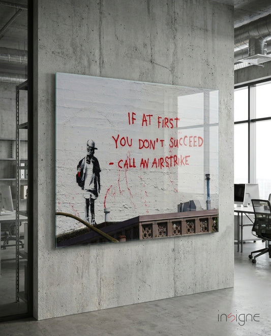 Glass art panel mounted on concrete wall in modern office, featuring stenciled child figure and red text reading "If at first you don't succeed - call an airstrike.