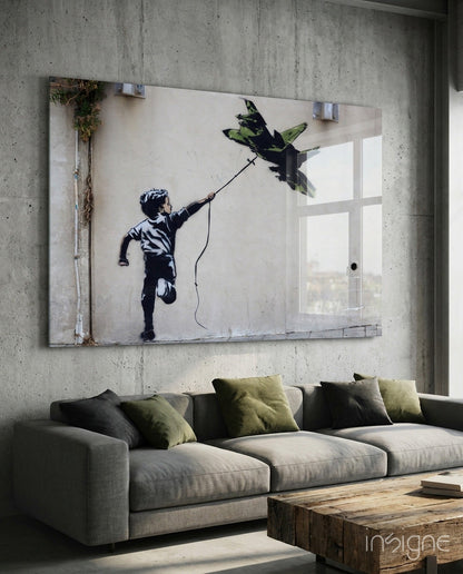 Large glass art panel mounted above gray sofa depicting stenciled silhouette of child flying fighter jet kite.