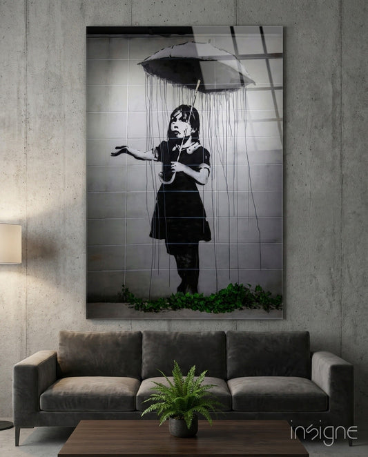 Black and white glass art depicting a young girl with umbrella in rain, displayed above gray sofa in concrete interior.