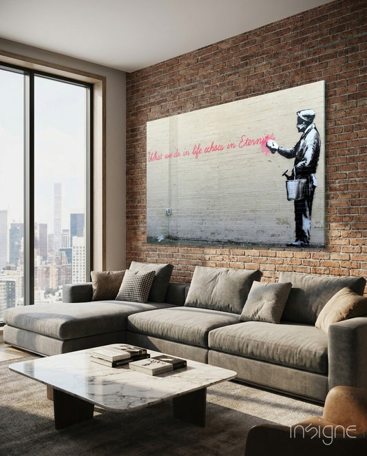 Banksy-style glass art print displayed above gray sectional sofa in modern loft with exposed brick and city views.