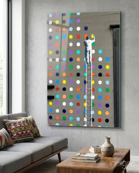 Artist on ladder installing colorful painted dots on large gray glass panel mounted on concrete wall above sofa.