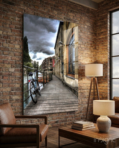 Glass wall art depicting a wooden boardwalk along a canal with a red bicycle and historic buildings under stormy sky.