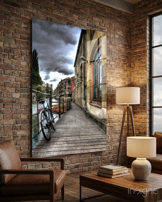 Glass wall art depicting a wooden boardwalk along a canal with a red bicycle and historic buildings under stormy sky.