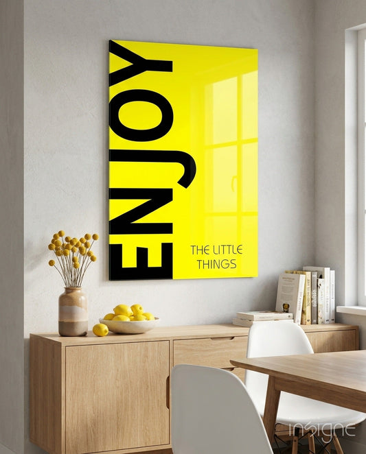 Bold yellow glass wall art with black "ENJOY THE LITTLE THINGS" typography displayed above wooden desk in minimalist interior.
