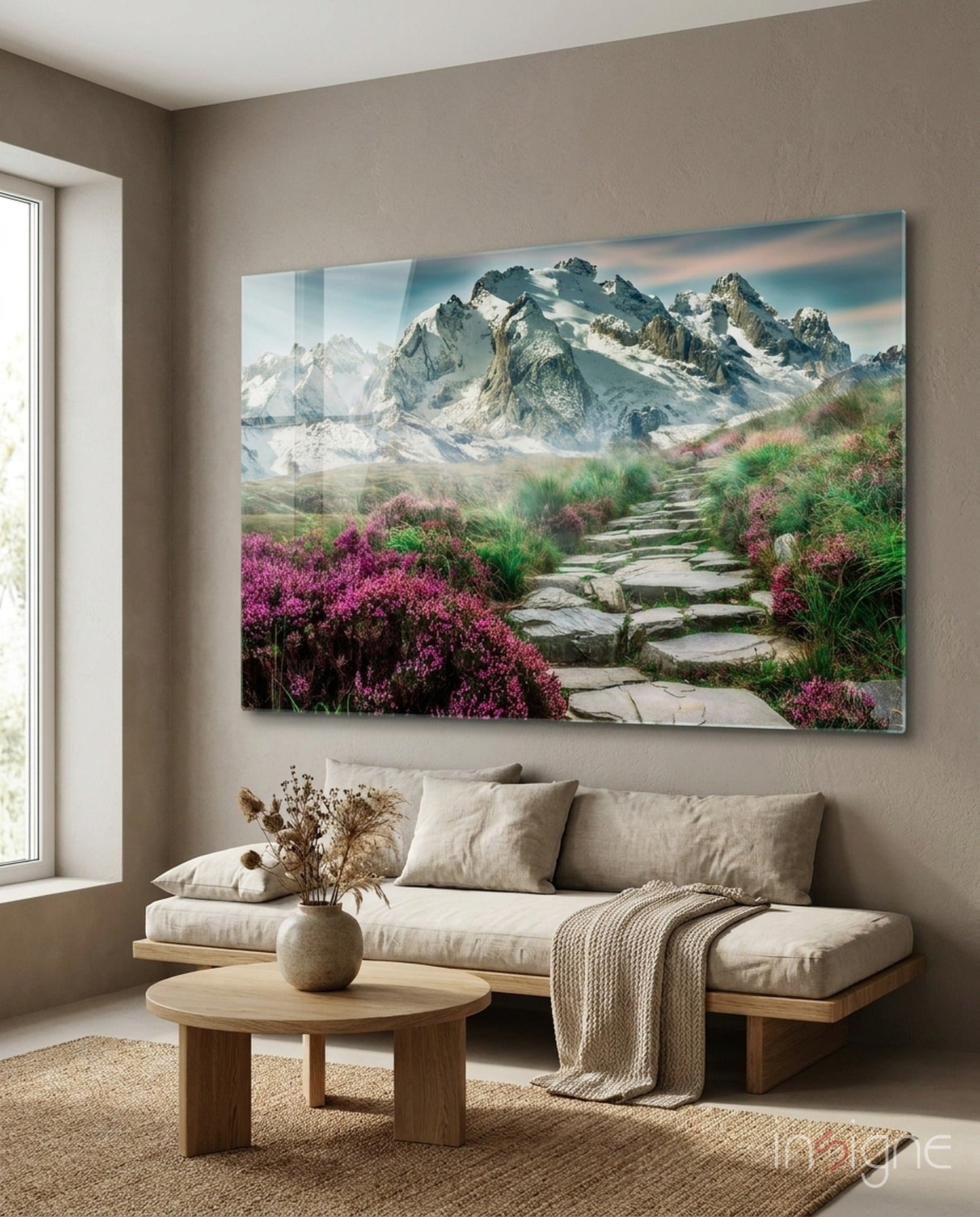 Glass wall art depicting a stone pathway through pink flowering shrubs leading to snow-capped mountains, hung above a beige sofa in a living room.