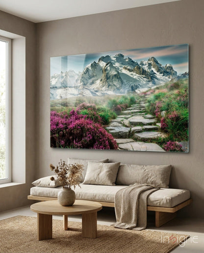 Glass wall art depicting a stone pathway through pink flowering shrubs leading to snow-capped mountains, hung above a beige sofa in a living room.
