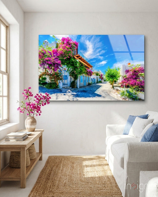 Aegean Beauties Glass Wall Art displayed above a white armchair in a bright room with magenta flowers and wooden furniture.