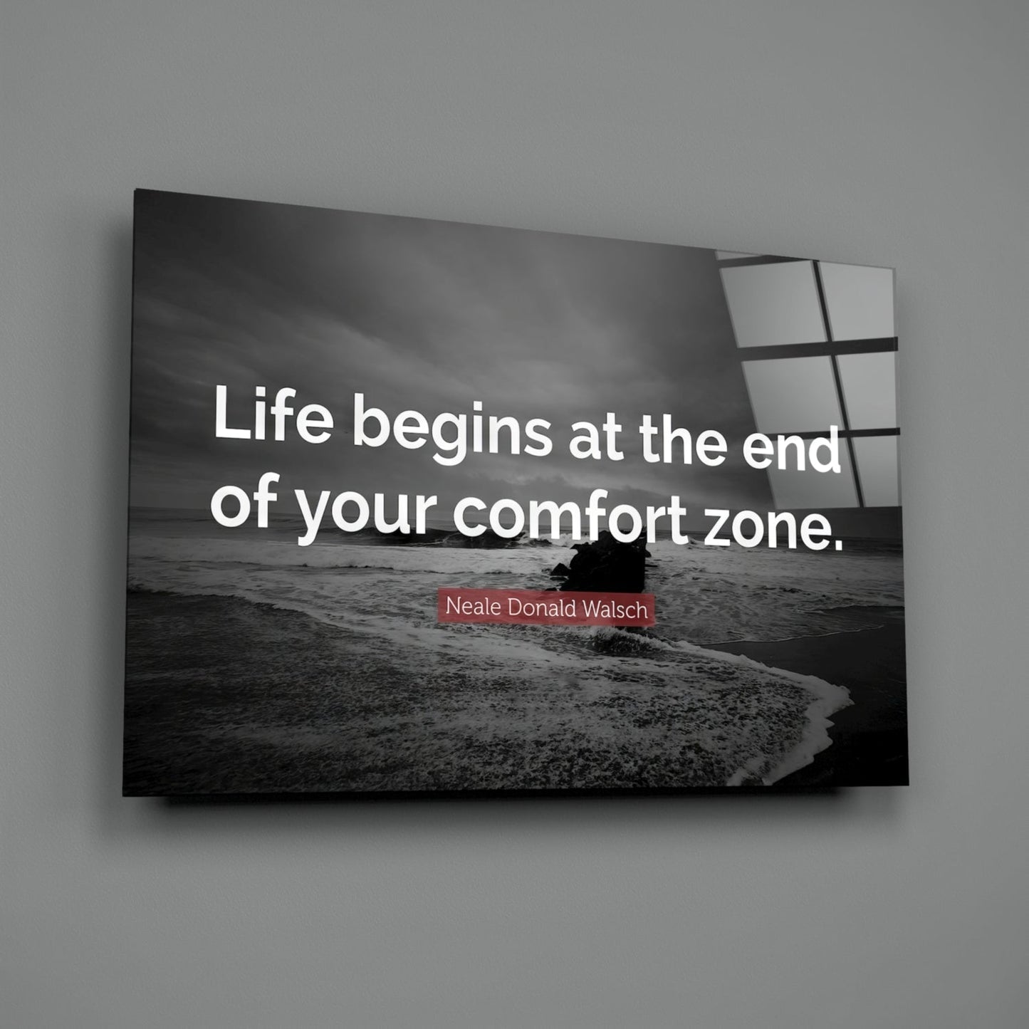 Black and white beach scene with waves and figure, displayed on glass wall art with white motivational text and red attribution to Neale Donald Walsch.