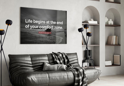 Modern living room interior with black leather sofa displaying glass wall art featuring quote "Life begins at the end of your comfort zone" above it.