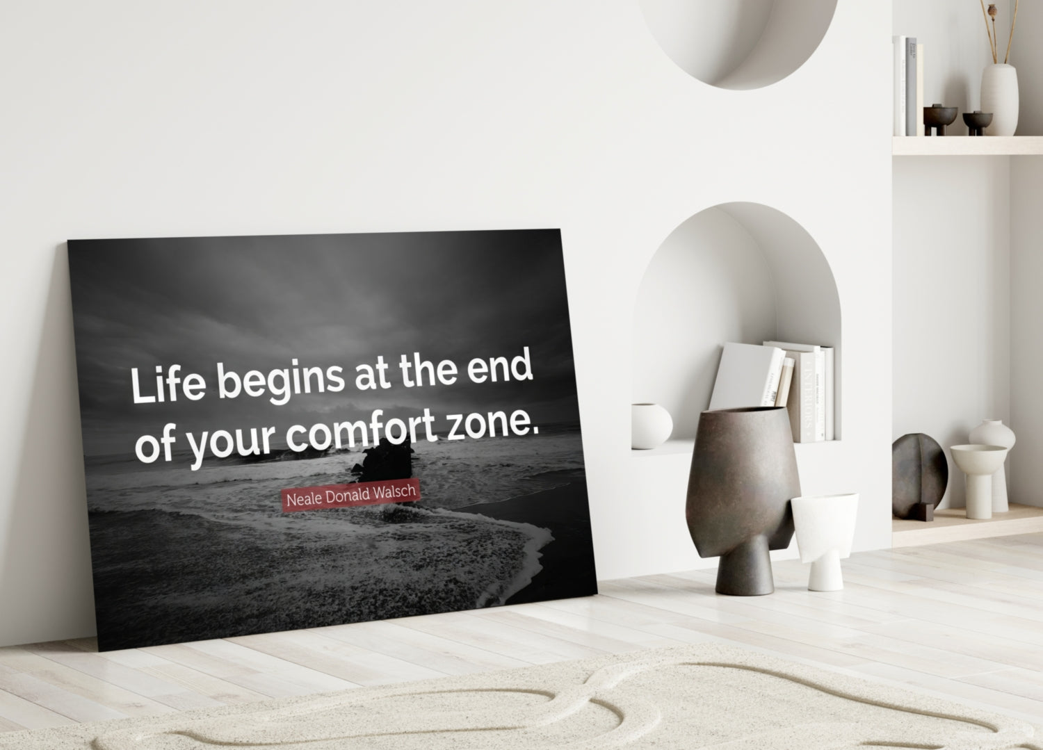 Black glass wall art with white text reading "Life begins at the end of your comfort zone" and attribution to Neale Donald Walsch, leaning against white interior wall with decorative objects.
