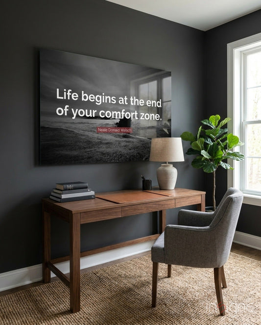 Black and white motivational glass wall art displaying "Life begins at the end of your comfort zone" quote above wooden desk in dark office.