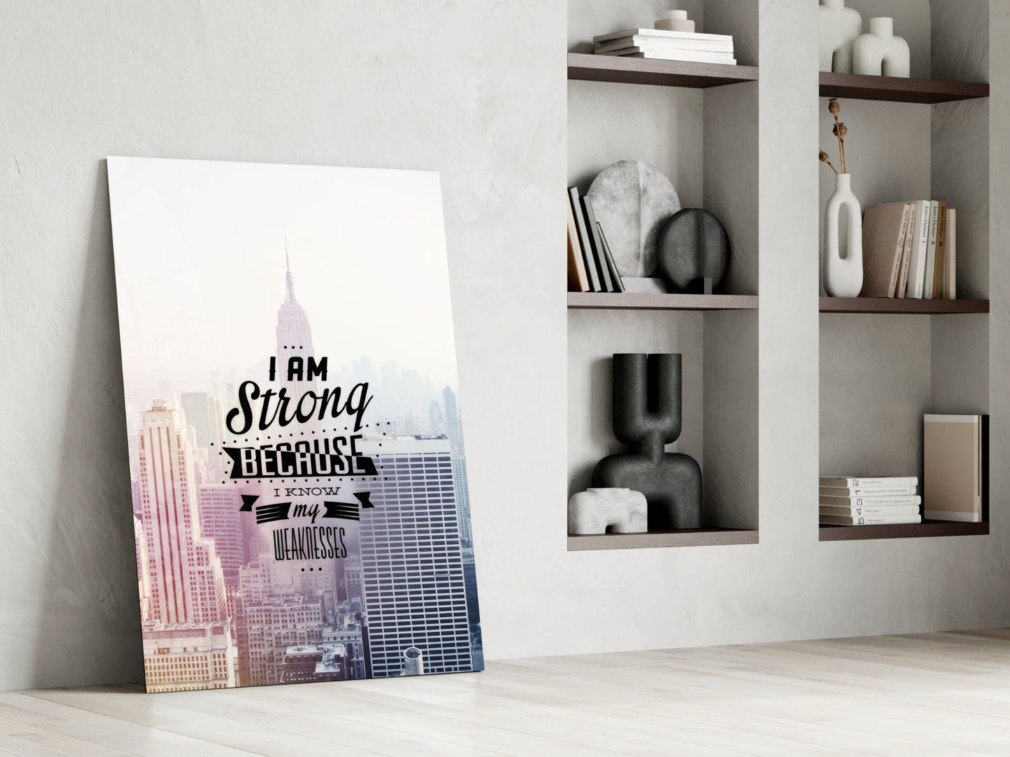 Glass wall art leaning against white wall beside shelving unit, featuring New York City skyline in pink and blue with black text reading "I am Strong Because I Know my Weaknesses.