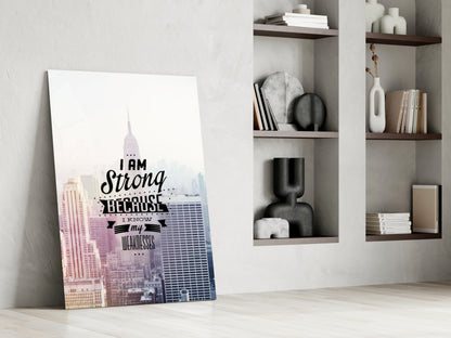 Glass wall art leaning against white wall beside shelving unit, featuring New York City skyline in pink and blue with black text reading "I am Strong Because I Know my Weaknesses.
