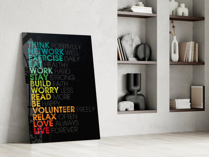 Black glass wall art with colorful gradient text displaying life advice phrases, leaning against white wall beside wooden shelving unit.
