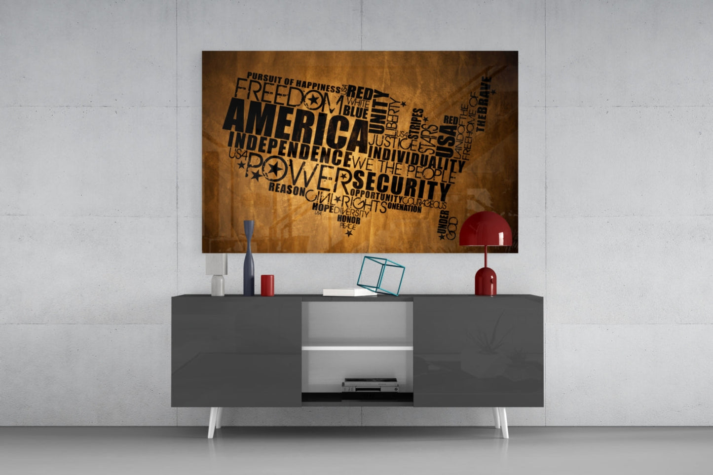 USA map word art glass wall panel in gold and black mounted above gray media console with red lamp and accessories.
