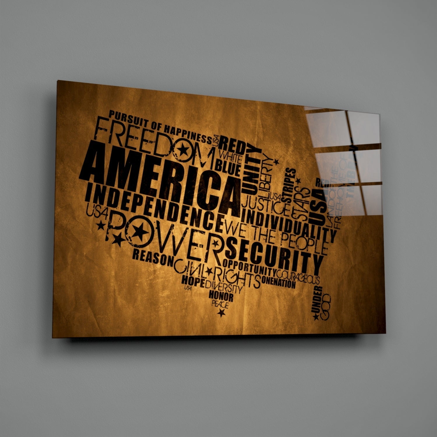 USA map-shaped glass wall art with patriotic words in black text on gold textured background, displayed at angle against gray wall.