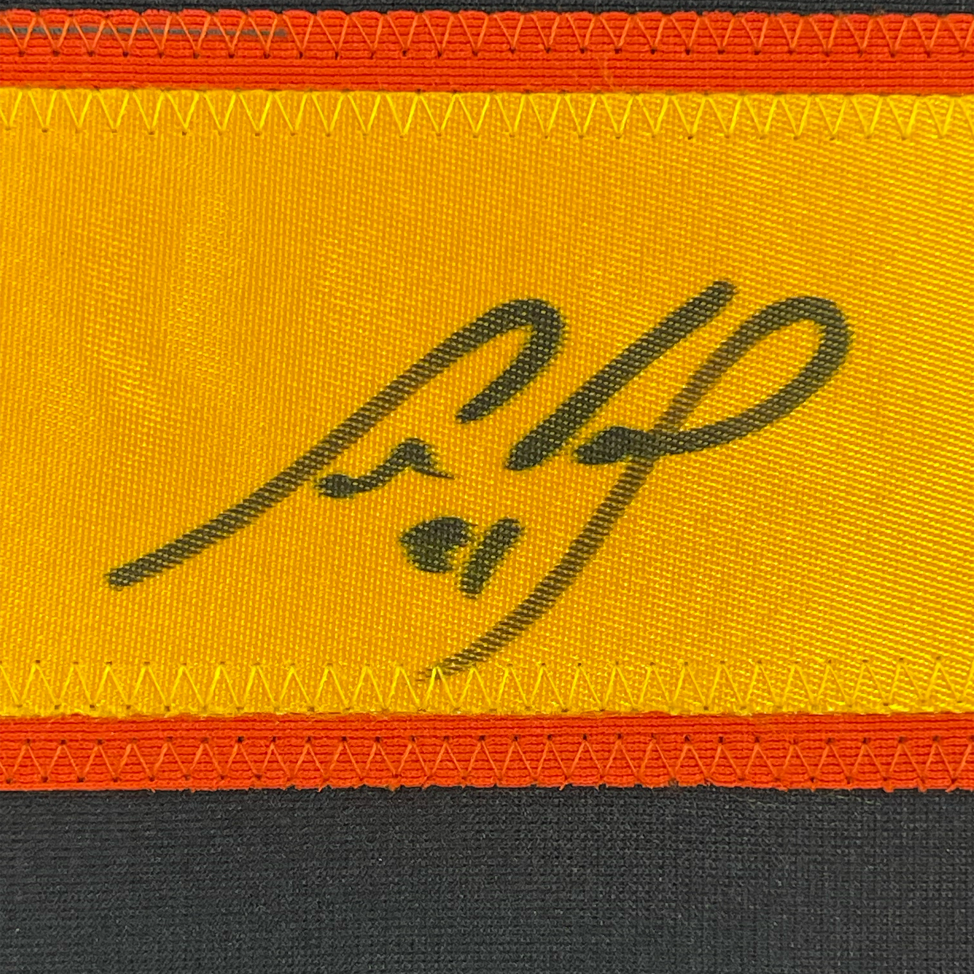 Close-up of signed autograph on yellow fabric strip with red and black borders, showing handwritten signature in dark ink.