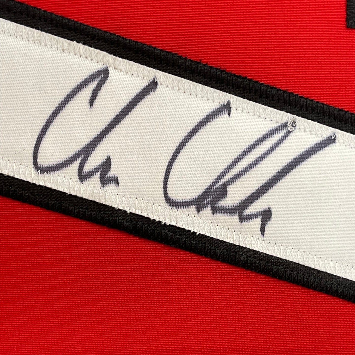 Close-up of autographed nameplate on white fabric strip with black borders against red jersey background.