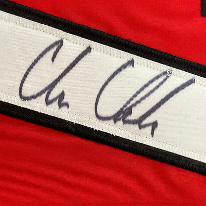 Close-up of autographed nameplate on white fabric strip with black borders against red jersey background.
