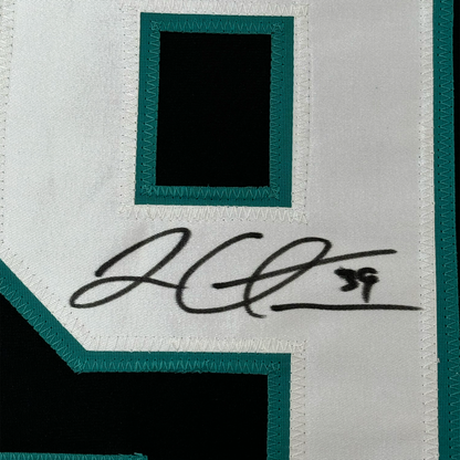 Close-up of black handwritten signature on white fabric with teal border matting and frame edge visible.