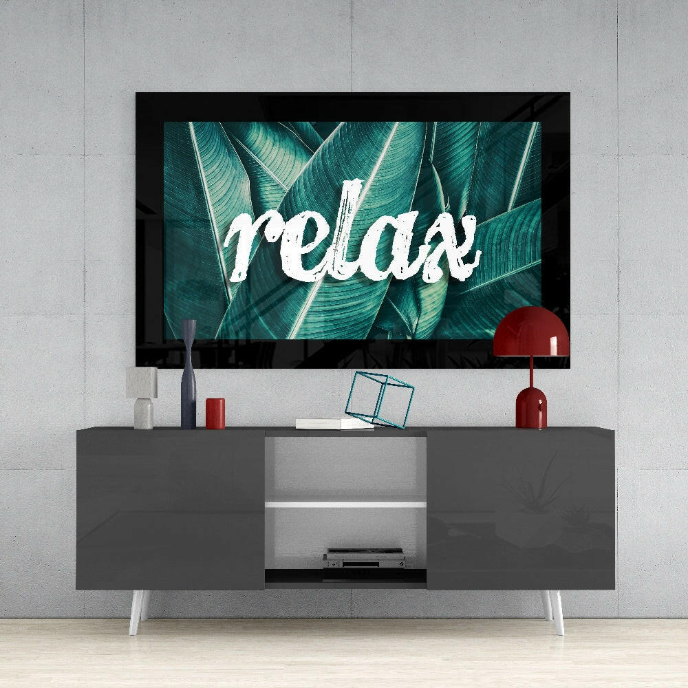Framed glass wall art displaying "relax" text over teal tropical leaves, mounted above a gray media console with red accents.