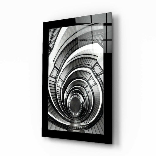 Black-framed glass wall art showing a dramatic downward view of a spiral staircase with concentric curved railings in black and white.