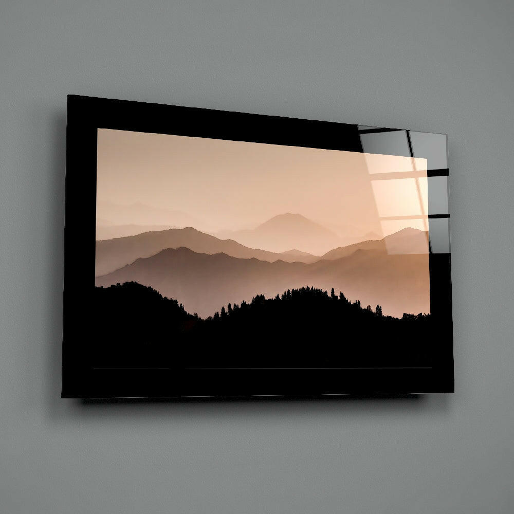 Black-framed glass wall art displaying layered misty mountains in warm beige tones with forested silhouettes, mounted on gray wall.