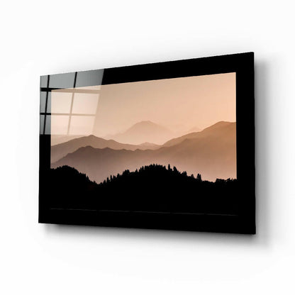 Black-framed glass wall art displaying layered mountain silhouettes against a warm peachy sky, shown at an angle.