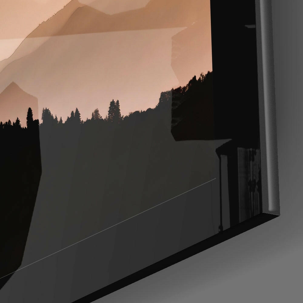 Glass wall art mounted on white wall displaying silhouetted forest treeline beneath misty peach-toned mountains.