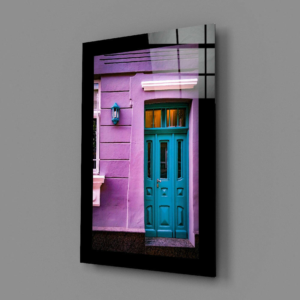 Black-framed glass wall art displaying a vibrant pink building facade with a turquoise door and cyan lantern, angled view.