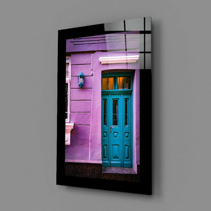 Black-framed glass wall art displaying a vibrant pink building facade with a turquoise door and cyan lantern, angled view.