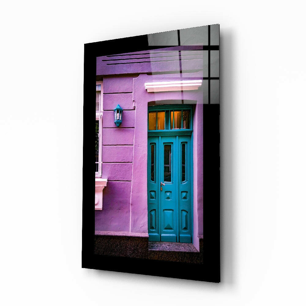 Glass wall art in black frame displaying vibrant pink building facade with turquoise door and window, angled perspective view.