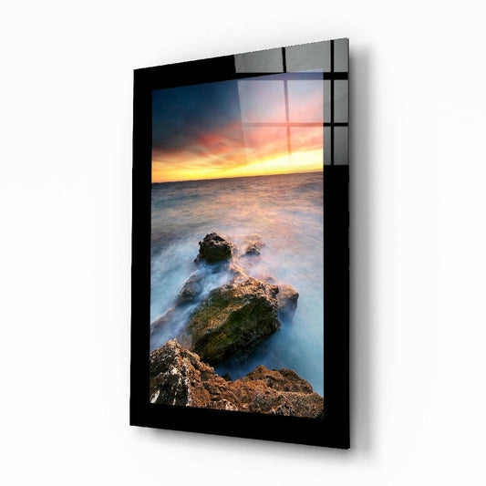 Angled product view of glass wall art featuring rocky coastline with golden sunset sky and misty ocean waves in black frame.