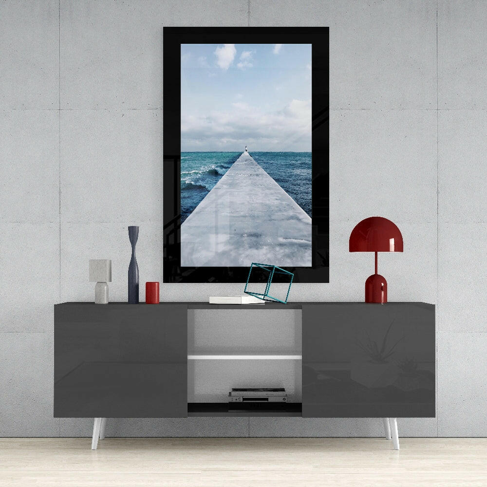 Black-framed glass wall art displaying a white pier extending into turquoise ocean beneath cloudy sky, mounted above gray media console.